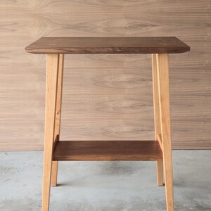 Puede incluir: Una peque&ntilde;a mesa auxiliar de madera con una parte superior rectangular y un estante inferior. La parte superior y el estante son de color marr&oacute;n oscuro, mientras que las patas son de un color madera natural m&aacute;s claro. La mesa mide unos 61 cm de altura.