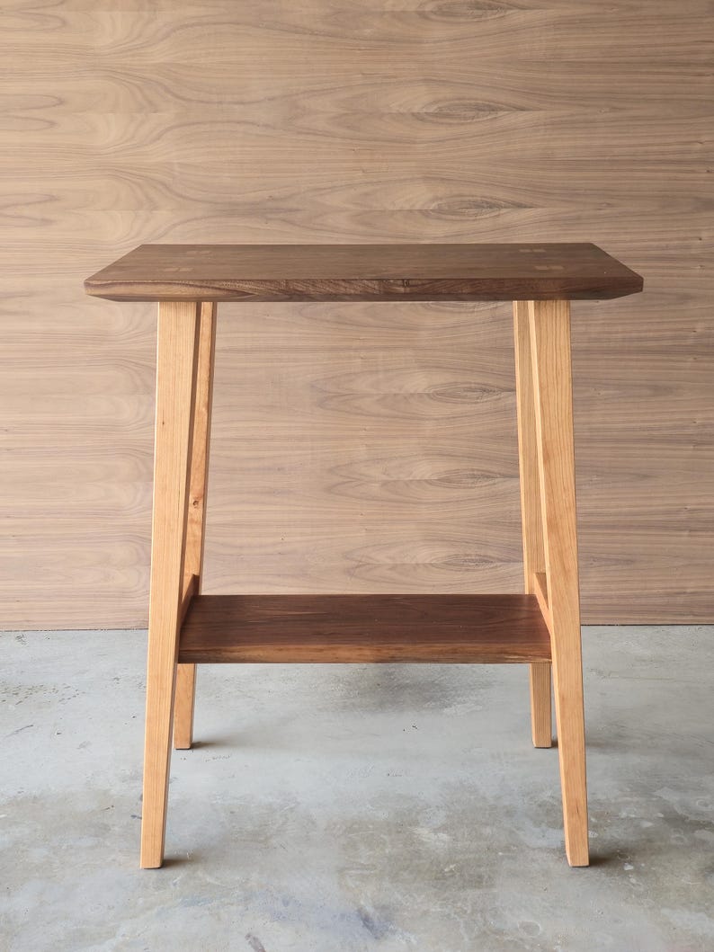 Puede incluir: Una peque&ntilde;a mesa auxiliar de madera con una parte superior rectangular y un estante inferior. La parte superior y el estante son de color marr&oacute;n oscuro, mientras que las patas son de un color madera natural m&aacute;s claro. La mesa mide unos 61 cm de altura.