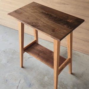 May include: A handcrafted wooden side table with a rectangular top and a lower shelf. The table features a dark brown tabletop with lighter wood legs and a shelf. The table is approximately 30 inches long, 12 inches wide, and 28 inches tall.