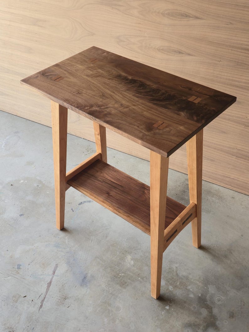 Puede incluir: Una mesa auxiliar de madera hecha a mano con una parte superior rectangular y un estante inferior. La mesa presenta una tapa marr&oacute;n oscuro con patas y un estante de madera m&aacute;s clara. La mesa mide aproximadamente 76 cm de largo, 30 cm de ancho y 71 cm de alto.