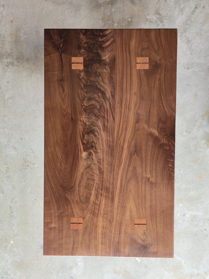 Puede incluir: Vista superior de una mesa rectangular de madera. La mesa es de madera marr&oacute;n oscura con un patr&oacute;n de vetas visible. Cuatro incrustaciones rectangulares de madera m&aacute;s clara est&aacute;n espaciadas uniformemente en la superficie, a&ntilde;adiendo un toque decorativo.