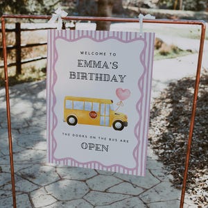 May include: A sign for Emma's birthday party, featuring a yellow school bus illustration with a pink heart balloon. The sign reads "Welcome to Emma's Birthday" and "The doors on the bus are open."