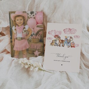 May include: A photograph of a young girl in a pink cowgirl outfit, and a greeting card with the text "Thank you". The card features a watercolor illustration of cartoon characters. The background is a soft, white fabric.