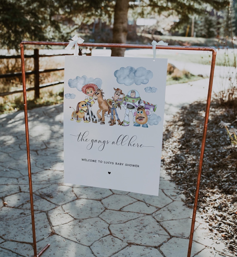 May include: A white sign featuring a watercolour illustration of Toy Story characters, including Woody and Buzz Lightyear. The sign reads "the gangs all here" and welcomes guests to Lucy's baby shower.