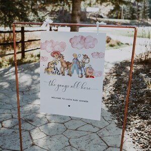 May include: A white sign with the text "the gangs all here" and "WELCOME TO LUCYS BABY SHOWER" is displayed on a copper-colored metal stand. The sign features watercolor illustrations of cartoon characters and pink clouds.
