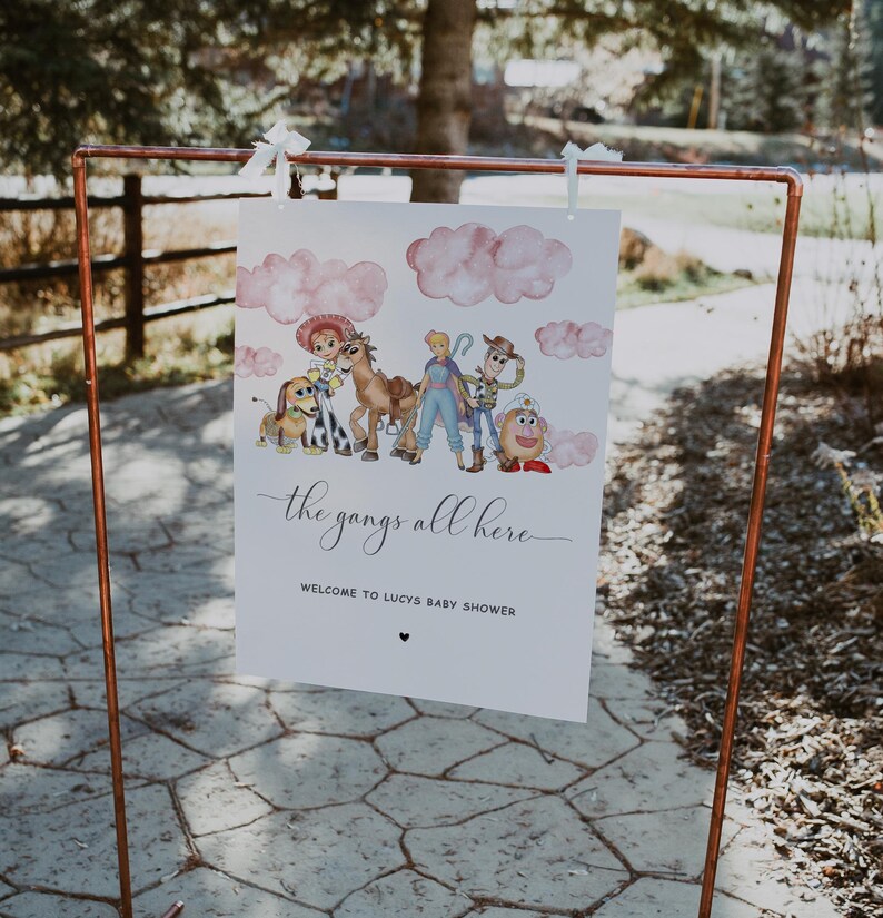 May include: A white sign with the text "the gangs all here" and "WELCOME TO LUCYS BABY SHOWER" is displayed on a copper-colored metal stand. The sign features watercolor illustrations of cartoon characters and pink clouds.