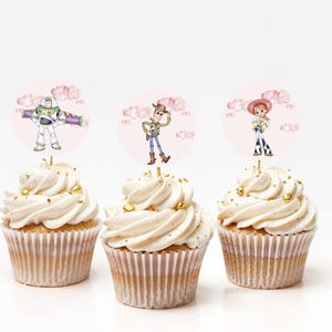 May include: Three cupcakes with white frosting and gold sprinkles. Each cupcake has a round topper featuring a character from the Toy Story movie: Buzz Lightyear, Woody, and Jessie. The toppers are set against a pink background with clouds.
