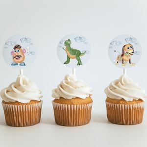May include: Three cupcakes with white icing and character toppers. The toppers feature illustrations of a cartoon potato, a green dinosaur, and a dachshund dog. The cupcakes are in brown paper cases.