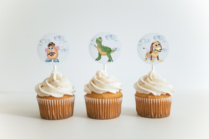 May include: Three cupcakes with white icing and character toppers. The toppers feature illustrations of a cartoon potato, a green dinosaur, and a dachshund dog. The cupcakes are in brown paper cases.