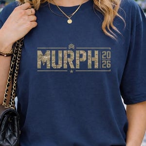 May include: Navy blue t-shirt with the word "MURPH" in a camouflage-style font, with the numbers "20" and "26" vertically aligned to the right. A star is centered below the word. The shirt is paired with blue jeans.
