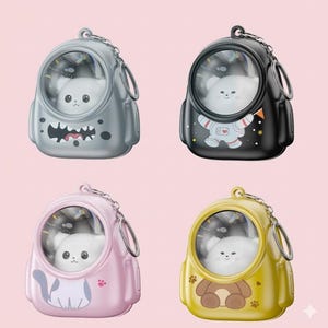 May include: Four keyrings shaped like astronaut helmets, each with a clear window showing a white plush animal. The keyrings are in grey, black, pink, and yellow. The black keyring has an astronaut graphic, and the pink one has a cat graphic.