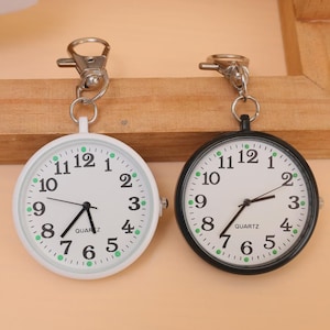 May include: Two pocket watches with quartz movements. One watch has a white case and the other has a black case. Both watches have white faces with black numerals and green dots. Each watch has a silver clip.