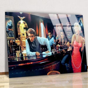 May include: A framed print featuring a retro bar scene with iconic figures. James Dean leans on the bar, while Elvis and Marilyn Monroe are also depicted. The bar has a jukebox and bottles.