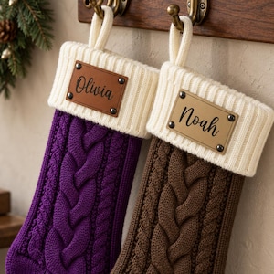 Puede incluir: Dos calcetines navideños de punto trenzado con puños acanalados de color crema. El calcetín morado tiene una etiqueta de aspecto cuero marrón con el nombre Olivia, y el marrón tiene una etiqueta beige con el nombre Noah, fijadas con remaches.
