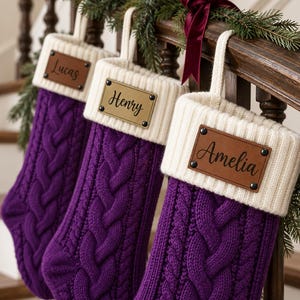 May include: Three purple cable-knit Christmas stockings with cream-coloured ribbed cuffs. Each stocking features a rectangular leather-like name tag attached with four black rivets, displaying the names Lucas, Henry, and Amelia in black script.