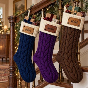 May include: Three cable-knit Christmas stockings in navy blue, purple, and brown. Each stocking features a cream-colored ribbed cuff with a personalized leather patch attached by metal rivets, displaying the names Julian, Amelia, and Mabel.