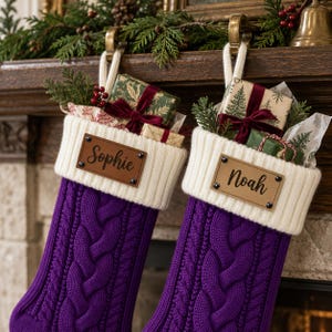 Puede incluir: Dos calcetines navideños de punto trenzado morado con puños acanalados blancos. Cada calcetín tiene un parche rectangular de cuero tostado sujeto con cuatro remaches negros. El nombre Sophie está grabado a la izquierda y Noah a la derecha.