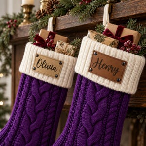 Könnte beinhalten: Zwei violette Weihnachtsstrümpfe aus Zopfstrick mit weißen Rippbündchen. Jeder Strumpf hat ein rechteckiges, lederartiges Namensschild, das mit vier Metallnieten befestigt ist und die Namen Olivia und Henry in schwarzer Schreibschrift zeigt.