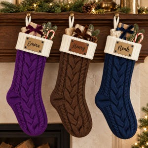 Könnte beinhalten: Drei Zopfstrick-Weihnachtsstrümpfe in Violett, Braun und Marineblau. Jeder Strumpf hat ein weißes Rippbündchen mit einem rechteckigen, lederartigen Namensschild mit der Aufschrift Emma, Henry oder Noah und ist mit einem kleinen Geschenk verziert.
