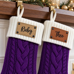 Könnte beinhalten: Zwei violette Weihnachtsstrümpfe aus Zopfstrick mit weißen Rippbündchen. Jeder Strumpf hat einen rechteckigen, hellbraunen Aufnäher aus Lederimitat, der mit vier dunklen Metallnieten befestigt ist. Links ist der Name Ruby und rechts Theo eingraviert.