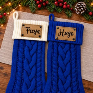 Puede incluir: Dos calcetines navideños de punto trenzado en azul real. El calcetín izquierdo tiene un puño acanalado blanco con una etiqueta de cuero marrón con el nombre Freya. El derecho tiene un puño azul con una etiqueta con el nombre Hugo.