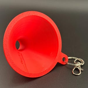 May include: A bright red funnel with a textured outer rim and a smooth interior surface. The funnel features a small tab with a silver-colored metal split ring and a lobster claw clasp attached, allowing it to function as a portable keychain accessory.