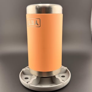 以下が含まれることがあります： オレンジ色の断熱タンブラー。銀色の蓋と「YETI」のロゴが付いています。タンブラーは、円形のデザインの灰色のベースにセットされています。背景は無地の濃い灰色です。