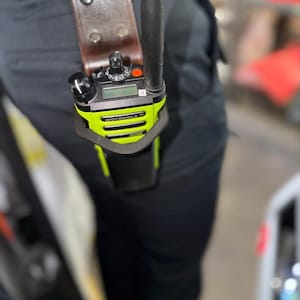 May include: A neon yellow and black Motorola two-way radio secured in a brown leather holster. The device features a prominent antenna, a small digital display, a circular volume knob, a channel selector dial, and a red button on the top panel.
