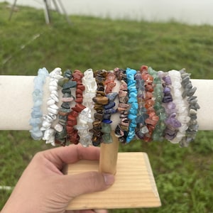Può includere: Una collezione di braccialetti con schegge di pietre preziose in vari colori, esposti su un supporto di legno. I braccialetti presentano pietre nelle tonalità di blu, bianco, rosso, marrone, verde e viola. Il supporto è tenuto da una mano su uno sfondo verde sfocato.