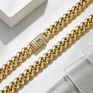 May include: A gold-toned cuban link chain necklace featuring a rectangular box clasp. The clasp is embellished with rows of clear baguette-cut stones and a central black square inlay containing the letter S in a gold-toned font.