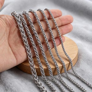 May include: Close-up of several silver-colored wheat chain necklaces. The chains are displayed on a wooden stand, with a soft gray background. The chains vary slightly in thickness, showcasing a range of styles for jewelry.