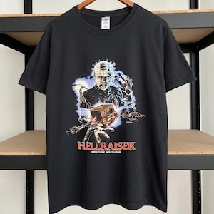 May include: A black short-sleeve t-shirt featuring a graphic print of Pinhead from Hellraiser. The design shows the character with chains and a puzzle box, with the red text HELLRAISER and the slogan DEMON TO SOME, ANGELS TO OTHERS below.