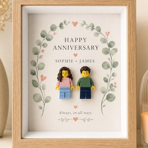 May include: A light wood shadow box frame containing a printed card with a green eucalyptus wreath, small pink hearts, and the text HAPPY ANNIVERSARY, SOPHIE + JAMES, and Always, in all ways. Two plastic toy figures are attached to the center.