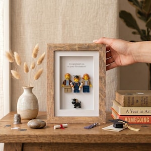 Puede incluir: Un marco de madera que contiene tres figuras de Lego y una pequeña figura de perro negro. La figura central lleva un birrete de graduación y sostiene un diploma. El texto "Congratulations on your Graduation!" aparece sobre un fondo blanco.