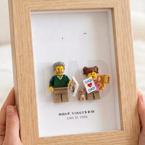 Puede incluir: Un marco de caja de madera clara con dos figuras LEGO y una pequeña figura de perro. La figura masculina sostiene un periódico, la femenina un trofeo dorado y un cartel que dice DAD CORAZÓN DAD. El texto dice HAPPY FATHER'S DAY JUNE 21, 2026.