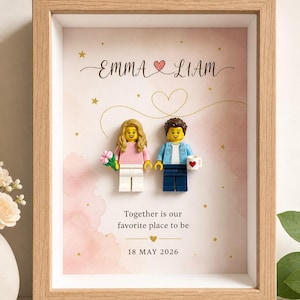 Puede incluir: Un marco de madera clara tipo caja con dos minifiguras de plástico. El fondo presenta un diseño de acuarela rosa con estrellas doradas, el texto "EMMA & LIAM", "Together is our favorite place to be" y la fecha "18 MAY 2026".