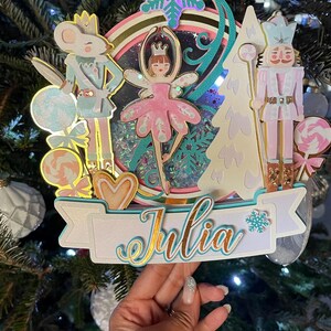 Peut inclure: Une décoration de gâteau multicouche représentant une ballerine centrale, un soldat souris et un soldat casse-noisette. Le design comprend un élément circulaire à paillettes bleues, un sapin blanc, des sucettes et une bannière avec le nom Julia en doré.