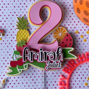 May include: A layered paper cake topper featuring a large pink number 2, the names Amirah Juliet in black and white script, and colorful fruit cutouts including a pineapple, strawberry, cherry, orange slice, and watermelon slice on a stick.