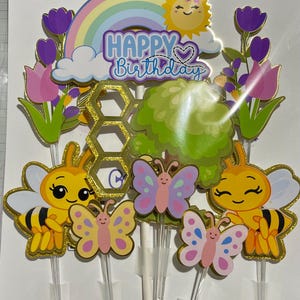 May include: A set of cake toppers featuring a rainbow, a smiling sun, and the text Happy Birthday. The set includes two yellow and black bees, three colourful butterflies, purple tulips, a green bush, and a gold glitter honeycomb shape.