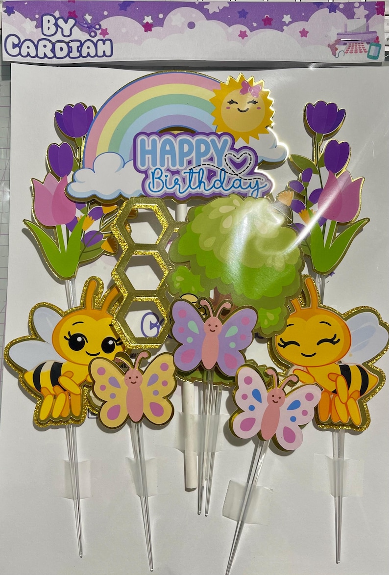 May include: A set of cake toppers featuring a rainbow, a smiling sun, and the text Happy Birthday. The set includes two yellow and black bees, three colourful butterflies, purple tulips, a green bush, and a gold glitter honeycomb shape.