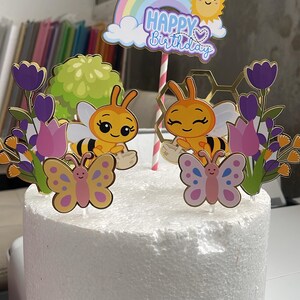 May include: A cake topper set featuring two cartoon bees, two butterflies, purple and pink tulips, a green bush, a gold honeycomb shape, and a rainbow arch with a sun and the text Happy Birthday, all mounted on sticks in a white foam cylinder.