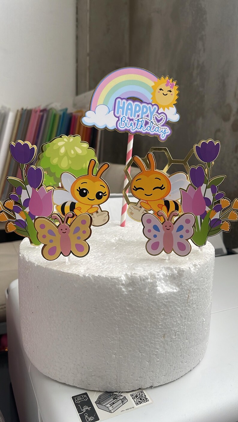 May include: A cake topper set featuring two cartoon bees, two butterflies, purple and pink tulips, a green bush, a gold honeycomb shape, and a rainbow arch with a sun and the text Happy Birthday, all mounted on sticks in a white foam cylinder.
