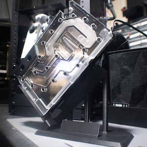 May include: A clear acrylic and black metal liquid cooling water block for a computer processor, featuring internal channels for coolant flow, mounted on a black plastic stand, with a black mesh power supply unit visible in the background.