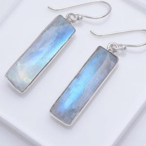 May include: A pair of rectangular earrings with a silver-colored frame. The earrings feature a light blue and white iridescent stone. The earrings have a hook closure. The jewelry is displayed on a white surface.