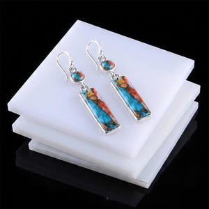 Puede incluir: Un par de pendientes colgantes de plata con incrustaciones rectangulares y circulares de piedra compuesta de turquesa y cobre multicolor. Los pendientes tienen un acabado de plata pulida y cuelgan de sencillos ganchos de plata para un ajuste seguro.