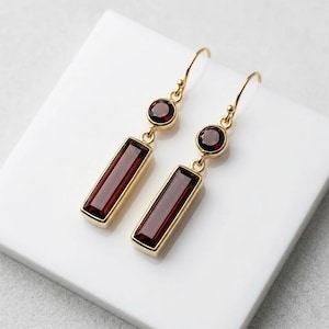 Può includere: Un paio di orecchini pendenti color oro con una gemma rettangolare rosso scuro e una gemma rotonda più piccola. Gli orecchini sono presentati su una superficie bianca. Le gemme hanno una superficie lucida e riflettente.