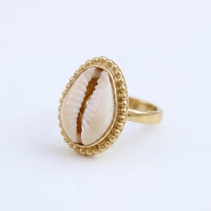 May include: A gold-colored ring featuring a cream-colored seashell centerpiece. The seashell is oval-shaped and set within a beaded gold frame. The ring band is a simple, polished gold. The ring is set against a white background.