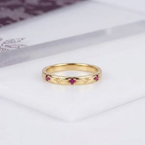 May include: A gold band ring featuring a series of alternating starburst engravings and small, round, deep pink gemstones set flush into the metal surface. The ring has a polished finish and a consistent width throughout its circular design.