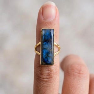 May include: A gold-toned ring featuring a rectangular, iridescent blue gemstone. The gemstone displays a range of blue hues. The ring's band has a delicate, open design. This piece of jewelry is a statement ring.