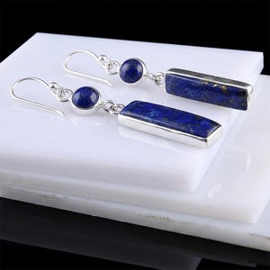 May include: A pair of silver earrings with a round, dark blue stone and a rectangular, dark blue stone. The earrings have a hook closure and are displayed on a white surface.